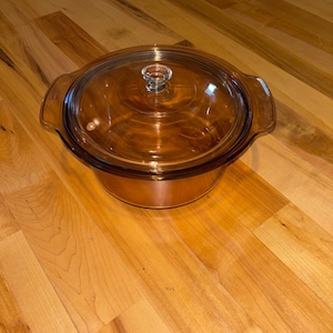 Puede incluir: Una fuente redonda de vidrio color ámbar con tapa a juego. La fuente tiene dos asas y un pomo de vidrio transparente en la tapa. El utensilio de cocina está diseñado para hornear y servir.