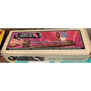 Puede incluir: Una caja de juego Gravitation vintage. La caja presenta el nombre del juego en una fuente estilizada con un fondo rosa y rojo. La caja también incluye el texto "Desafía la ley de la gravedad de Newton... Haz que la bola de acero ruede cuesta arriba para anotar."