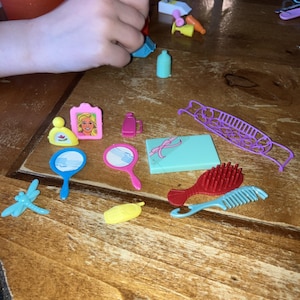 Op de afbeelding: Een verzameling miniatuur poppenaccessoires, waaronder een roze ingelijste foto, twee handspiegels, een blauwe libel, een rode haarborstel en een lichtblauwe kam. Andere items zijn een kleine parfumfles, een geschenkdoos en een paars decoratief stuk.