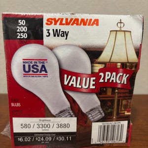 May include: A Sylvania 3-way light bulb 2-pack box featuring two white A21 bulbs. The packaging displays the text Made in the USA, Value 2 Pack, and brightness levels of 580, 3300, and 3880 lumens. A lamp illustration is printed on the right side.