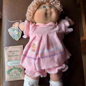 Könnte beinhalten: Cabbage Patch Kids Puppe mit blonden Garnhaaren, trägt ein rosa Kleid und passende Shorts. Die Puppe hat ein lächelndes Gesicht und trägt weiße Schuhe. Eine Geburtsurkunde und Adoptionspapiere liegen neben der Puppe.
