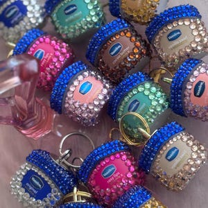 May include: A collection of bedazzled Vaseline lip therapy containers with blue lids and various jewel tones. Flavors include cocoa butter, rosy lips, and vanilla swirl. Each container has a silver keychain attached.