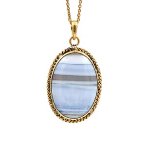 May include: An oval pendant necklace featuring a banded blue and grey stone set in a gold-toned twisted rope frame. The pendant hangs from a delicate gold-toned chain, showcasing horizontal layers of light blue, white, and dark grey within the stone.