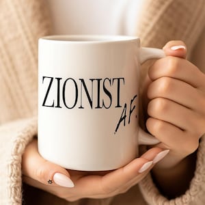 May include: White ceramic mug with the text "ZIONIST AF" in black. The mug is held by a person wearing a beige sweater. The mug has a handle on the right side.