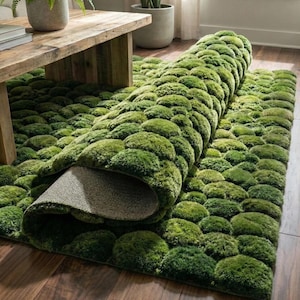 May include: A green rug with a textured, moss-like design. The rug is partially rolled up, revealing a gray backing. The rug's surface is covered in rounded, tufted shapes in varying shades of green, creating a three-dimensional effect. The rug is on a wooden floor.