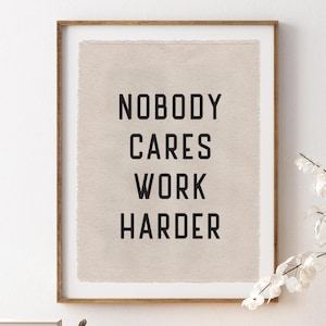 May include: A framed print with the text "NOBODY CARES WORK HARDER" in black letters. The print is displayed above a wooden dresser with a woven basket and a vase of white flowers.