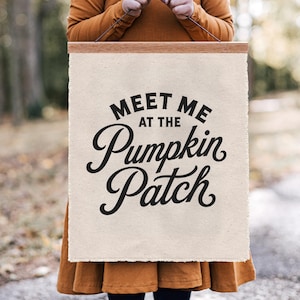 May include: A rectangular off-white canvas wall hanging featuring the black printed text "MEET ME AT THE PUMPKIN PATCH" in a mix of serif and script fonts. The canvas is secured at the top by a light brown wooden frame with a thin hanging cord.