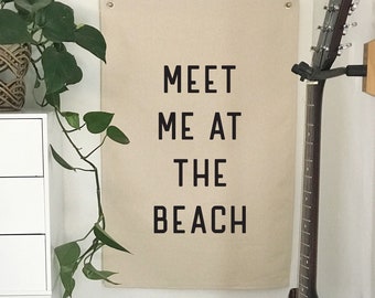 May include: A room corner featuring a canvas wall hanging with the text "MEET ME AT THE BEACH" in black. A dark guitar is mounted on the wall, and a black amplifier sits below. A white cabinet with black label holders and a potted plant complete the scene.