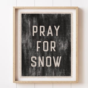 May include: A framed print with the words "PRAY FOR SNOW" in a bold, white font against a textured black background. The print is in a light-colored wooden frame, suitable for wall decor.