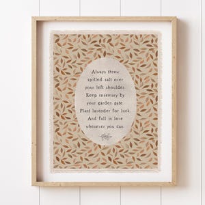 May include: A framed print with a beige background featuring a repeating pattern of brown leaves. The print includes an oval-shaped text area with the words "Always throw spilled salt over your left shoulder..." in a serif font.