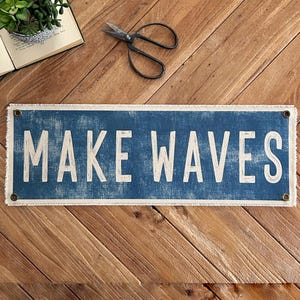 May include: A rectangular blue sign with the words "MAKE WAVES" in white lettering. The sign has a distressed look with a frayed white border and is mounted on a wooden surface. A pair of scissors and a small potted plant are also visible.
