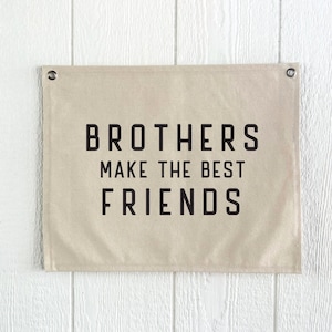 May include: A rectangular beige banner with the text "BROTHERS MAKE THE BEST FRIENDS" in black block letters. The banner has metal grommets in the top corners and is hanging against a white wooden wall.
