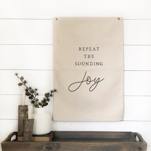 May include: A rectangular beige canvas wall hanging featuring the black printed text "REPEAT THE SOUNDING JOY" with two metal grommets at the top corners for mounting. The fabric banner is displayed vertically against a white horizontal plank wall.