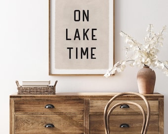 May include: A framed print with the words "ON LAKE TIME" in black block letters. The print hangs above a wooden dresser with a woven basket and a vase of white flowers. A wooden chair sits in front of the dresser.