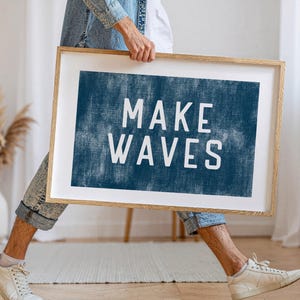 May include: A framed art print with the words "MAKE WAVES" in white block letters on a textured navy blue background. The print is in a light wood frame. The image is held by a person.