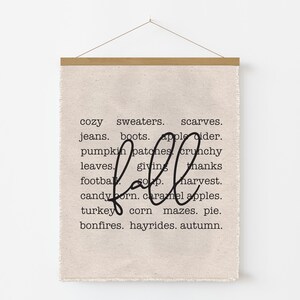 May include: A beige fabric wall hanging with a wooden bar at the top and a string for hanging. The word "fall" is written in a cursive font, with a list of fall-themed words in a block font.