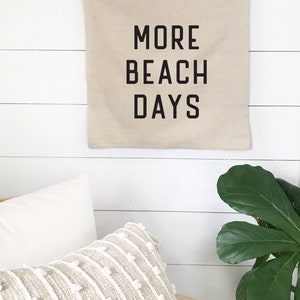 Puede incluir: Un banner de pared de lona beige con texto negro que dice "MORE BEACH DAYS". El banner est&aacute; colgado en una pared de madera blanca.