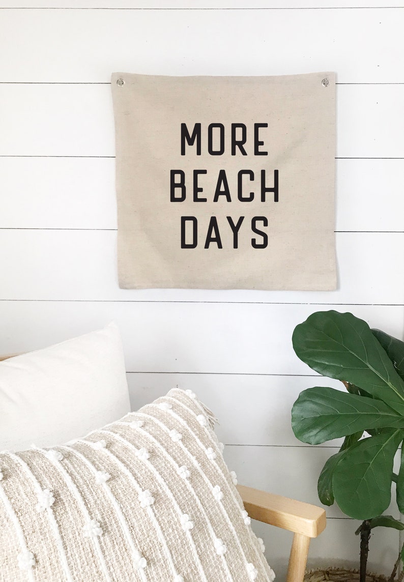 Puede incluir: Un banner de pared de lona beige con texto negro que dice "MORE BEACH DAYS". El banner est&aacute; colgado en una pared de madera blanca.