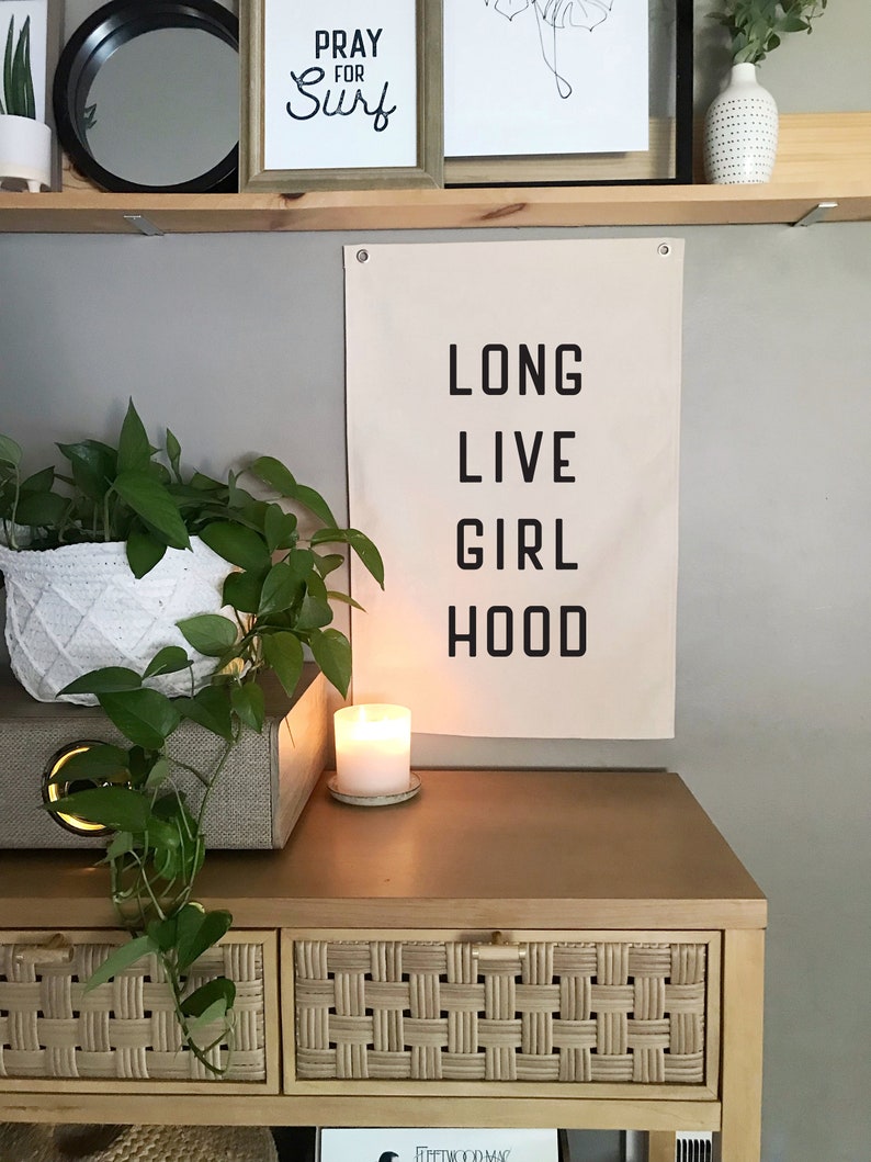 以下が含まれることがあります： 「LONG LIVE GIRL HOOD」の文字が黒いブロック体で書かれたベージュ色のキャンバスバナーが壁に掛けられています。 燃えているろうそくが木製の表面にあり、その下に編まれたバスケットの引き出しがあります。 装飾品が上の棚にあります。
