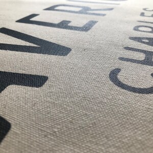 以下が含まれることがあります： 「RIVER」と「CHARLES」という名前が濃いグレーで印刷されたキャンバスのクローズアップ。文字は大きく太字で、少し使い古されたように見えます。キャンバスはニュートラルな薄いベージュ色です。