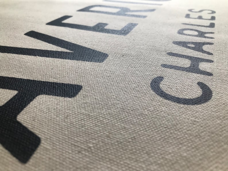 以下が含まれることがあります： 「RIVER」と「CHARLES」という名前が濃いグレーで印刷されたキャンバスのクローズアップ。文字は大きく太字で、少し使い古されたように見えます。キャンバスはニュートラルな薄いベージュ色です。