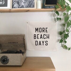 Puede incluir: Un tapiz de pared de lona beige con texto negro que dice "MORE BEACH DAYS". El tapiz est&aacute; colgado en una pared blanca sobre un armario de madera con cajones de rat&aacute;n tejido. Un tocadiscos vintage est&aacute; encima del armario.