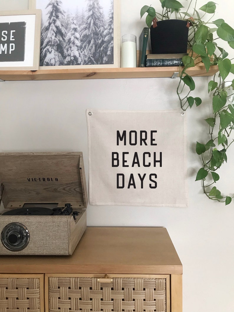 Puede incluir: Un tapiz de pared de lona beige con texto negro que dice "MORE BEACH DAYS". El tapiz est&aacute; colgado en una pared blanca sobre un armario de madera con cajones de rat&aacute;n tejido. Un tocadiscos vintage est&aacute; encima del armario.