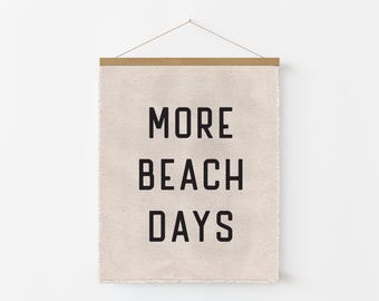 May include: A beige fabric wall hanging with the words "MORE BEACH DAYS" in bold black letters. The top edge is held by a wooden bar, and the bottom edge has a frayed finish. A simple string is used for hanging.