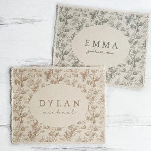 May include: Two beige fabric squares with a toile pattern and a white oval with a name and middle name printed in brown. The top square has the name "Emma June" and the bottom square has the name "Dylan Michael".