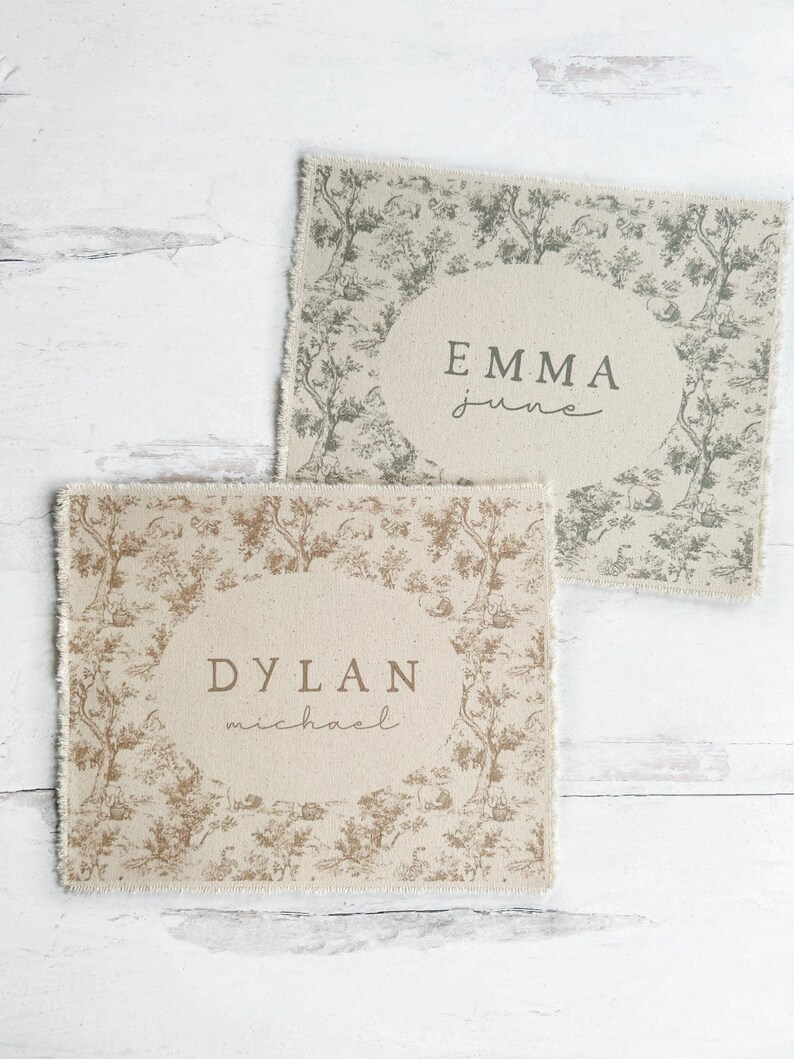 May include: Two beige fabric squares with a toile pattern and a white oval with a name and middle name printed in brown. The top square has the name "Emma June" and the bottom square has the name "Dylan Michael".