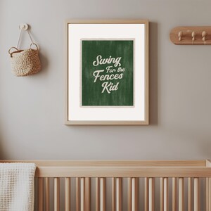 May include: A framed print with the text "Swing for the Fences Kid" in white script on a green background. The print is in a light wood frame and is hanging on a grey wall. A woven basket and a cot are also visible.
