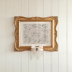 May include: A gold framed picture with a white background and a green floral pattern. The name "Emma June" is written in the center of the picture.