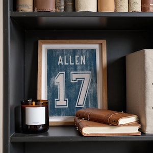 May include: A framed print of a blue jersey with the name "ALLEN" and the number "17" in white. A brown leather journal and a lit candle with a white label sit on a dark shelf. A beige storage box is also on the shelf.