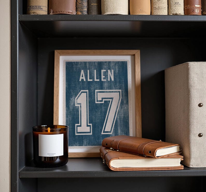 May include: A framed print of a blue jersey with the name "ALLEN" and the number "17" in white. A brown leather journal and a lit candle with a white label sit on a dark shelf. A beige storage box is also on the shelf.