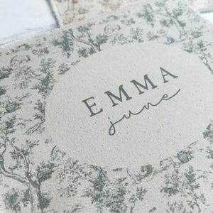 May include: A beige fabric with a green toile pattern and the name "EMMA june" printed in black.