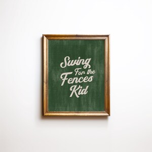 May include: A framed print with the text "Swing for the Fences Kid" in a white, cursive font. The background is a textured green, and the frame is a warm brown colour. The print is centered on a white background.