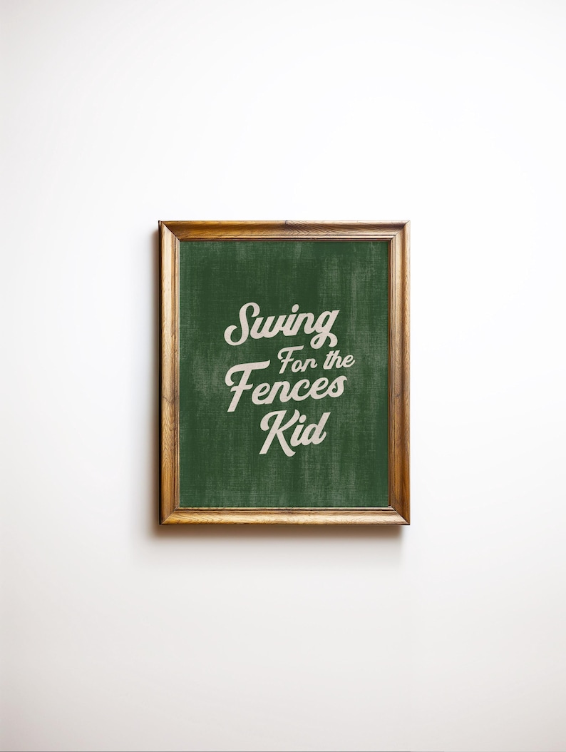 May include: A framed print with the text "Swing for the Fences Kid" in a white, cursive font. The background is a textured green, and the frame is a warm brown colour. The print is centered on a white background.