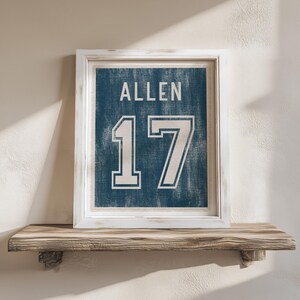 May include: Framed wall art featuring a blue background with the name "ALLEN" and the number "17" in white. The artwork is displayed in a white frame and rests on a wooden shelf.