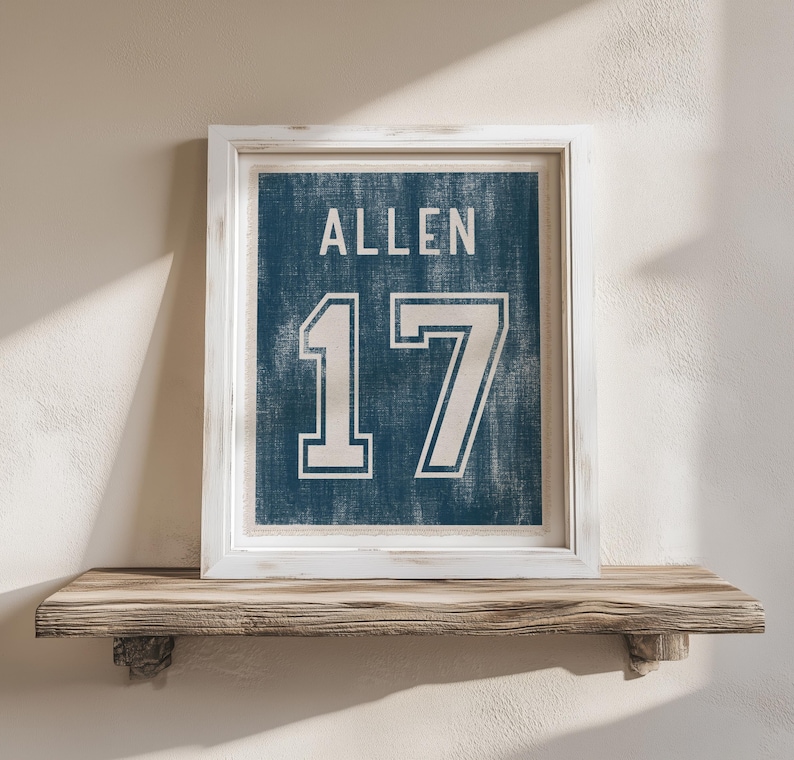 May include: Framed wall art featuring a blue background with the name "ALLEN" and the number "17" in white. The artwork is displayed in a white frame and rests on a wooden shelf.