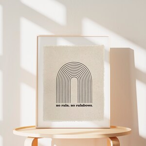 May include: A framed print with a black and white minimalist rainbow design and the text "no rain, no rainbows." The print is on a beige background and is displayed on a light wood table.