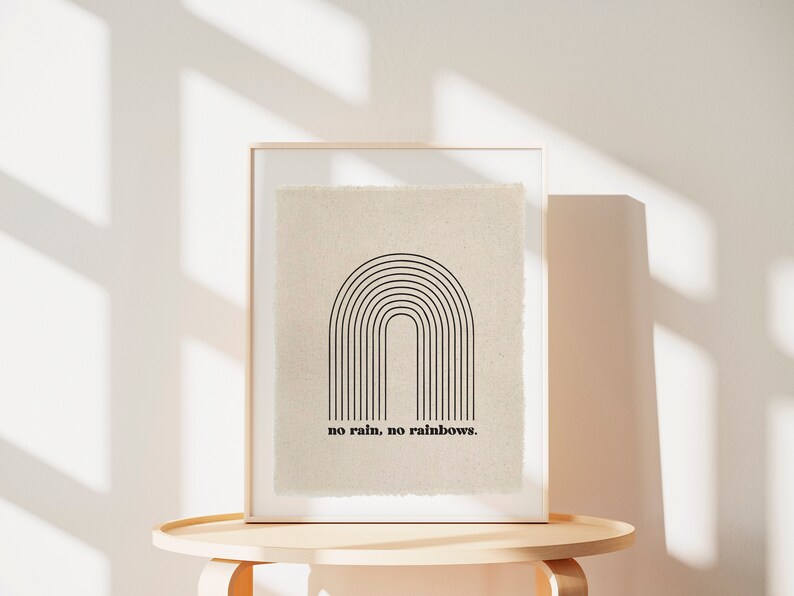 May include: A framed print with a black and white minimalist rainbow design and the text "no rain, no rainbows." The print is on a beige background and is displayed on a light wood table.