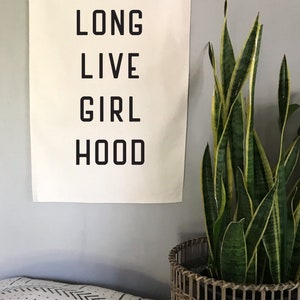以下が含まれることがあります： 「LONG LIVE GIRL HOOD」という言葉が黒い太字で書かれたクリーム色の壁掛け。バナーは灰色の壁に掛けられており、編まれたバスケットに入った鉢植えのサンスベリアの隣にあります。模様入りのクッションが手前にあります。