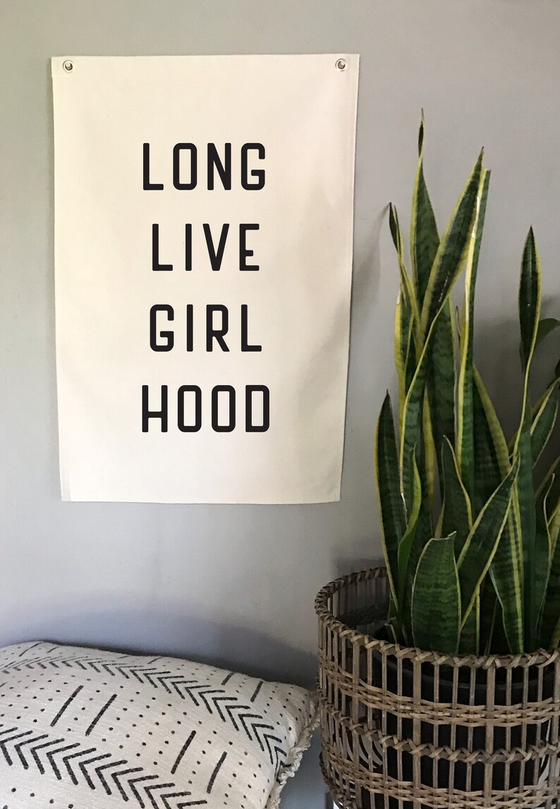 以下が含まれることがあります： 「LONG LIVE GIRL HOOD」という言葉が黒い太字で書かれたクリーム色の壁掛け。バナーは灰色の壁に掛けられており、編まれたバスケットに入った鉢植えのサンスベリアの隣にあります。模様入りのクッションが手前にあります。