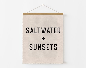 May include: A canvas wall hanging with the words "SALTWATER + SUNSETS" in bold, black letters. The canvas is a light beige color, and the top is held by a wooden bar with a string for hanging. The overall theme is coastal decor.