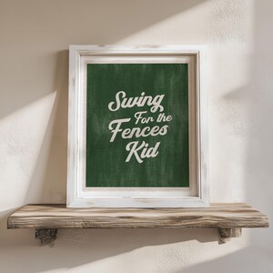 May include: A framed print with the text "Swing for the Fences Kid" in white script on a dark green background. The frame is white, and the print rests on a wooden shelf.