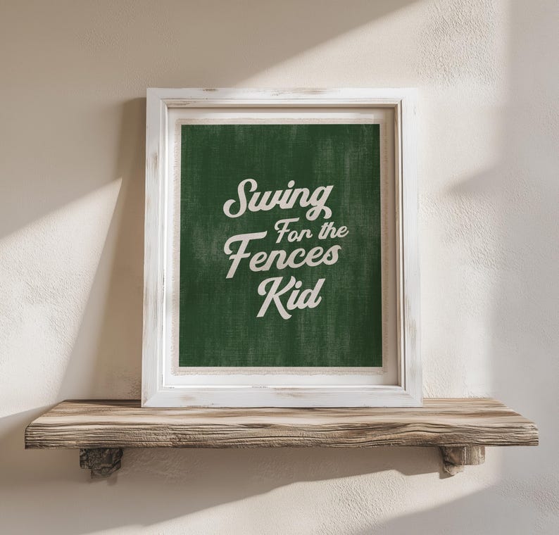 May include: A framed print with the text "Swing for the Fences Kid" in white script on a dark green background. The frame is white, and the print rests on a wooden shelf.