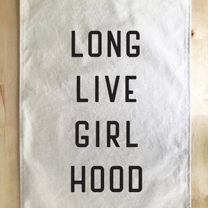 以下が含まれることがあります： 「LONG LIVE GIRL HOOD」の文字が太字の黒い大文字でプリントされた、長方形のオフホワイトのバナー。バナーには、吊り下げ用の金属グロメットが上隅にあります。背景は明るい木目です。