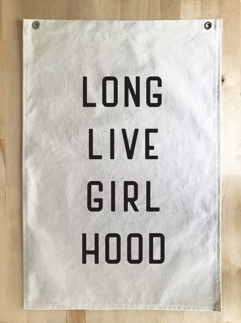 以下が含まれることがあります： 「LONG LIVE GIRL HOOD」の文字が太字の黒い大文字でプリントされた、長方形のオフホワイトのバナー。バナーには、吊り下げ用の金属グロメットが上隅にあります。背景は明るい木目です。