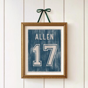 May include: Framed wall art featuring a blue jersey design with the name "ALLEN" and the number "17" in white. The artwork is in a gold frame and is suspended by a green ribbon against a white background.
