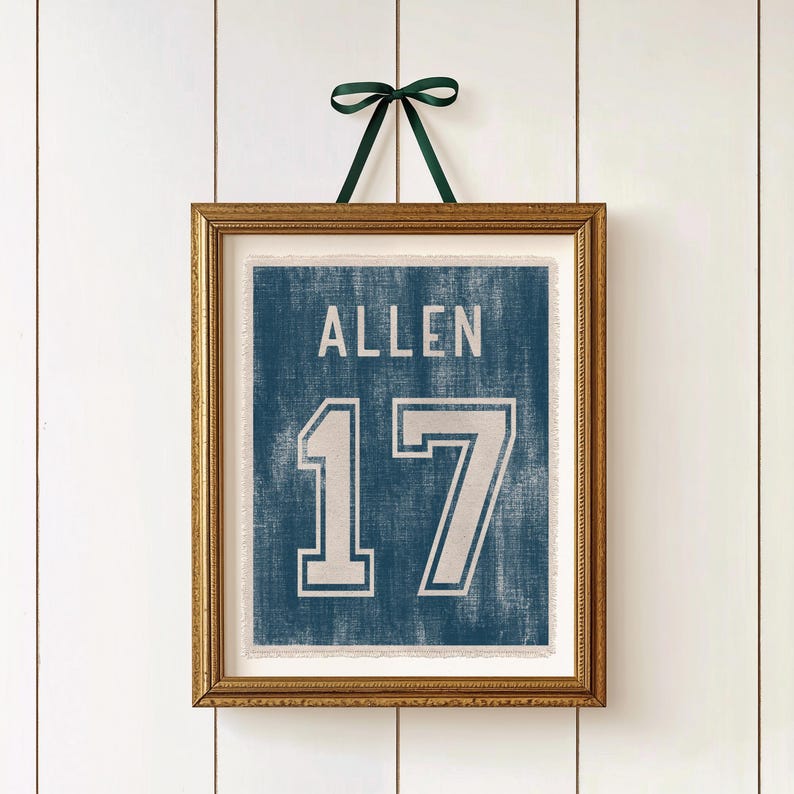 May include: Framed wall art featuring a blue jersey design with the name "ALLEN" and the number "17" in white. The artwork is in a gold frame and is suspended by a green ribbon against a white background.
